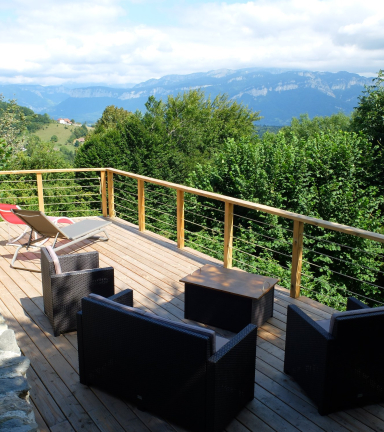 Une terrasse en bois est partage entre l'ombre et la lumire. Au soleil, deux chaises longues sont orientes vers la montagne.  l'ombre, des fauteuils et un canap entourent une table basse.
